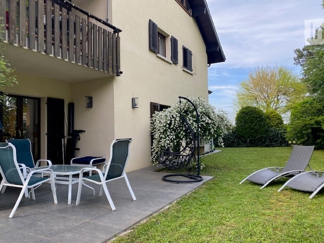 Outdoor patio area with a small seating set, including two chairs and a table, adjacent to a house with a balcony. A hanging chair is positioned near a flowering bush, and a lounge chair is placed on the lawn.