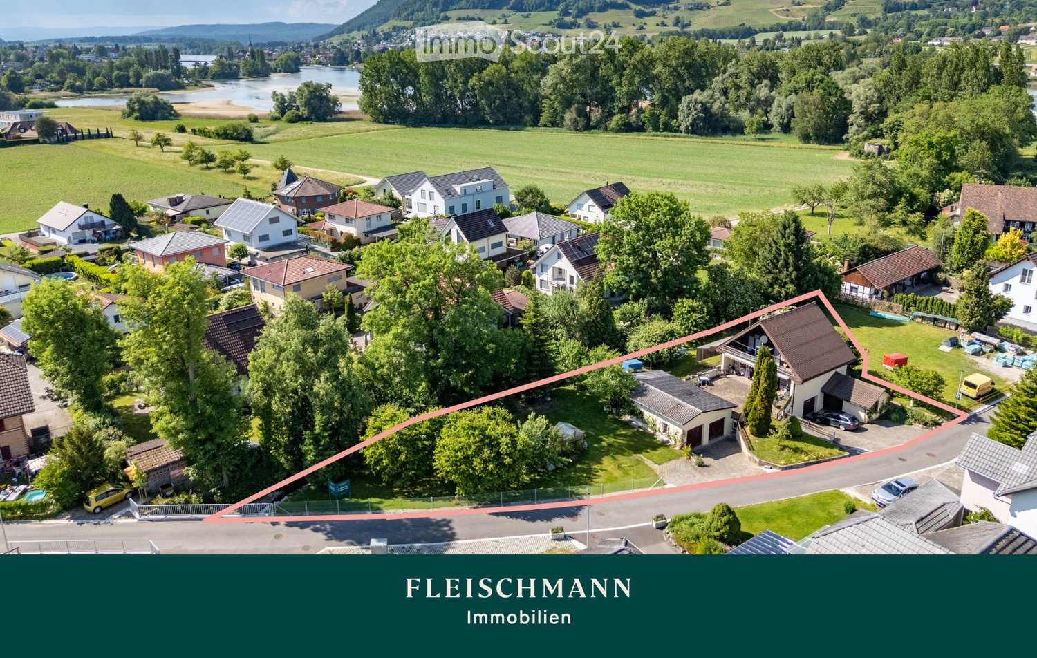 Aerial view of a residential area with multiple houses, surrounded by green fields, trees, and a river in the background.