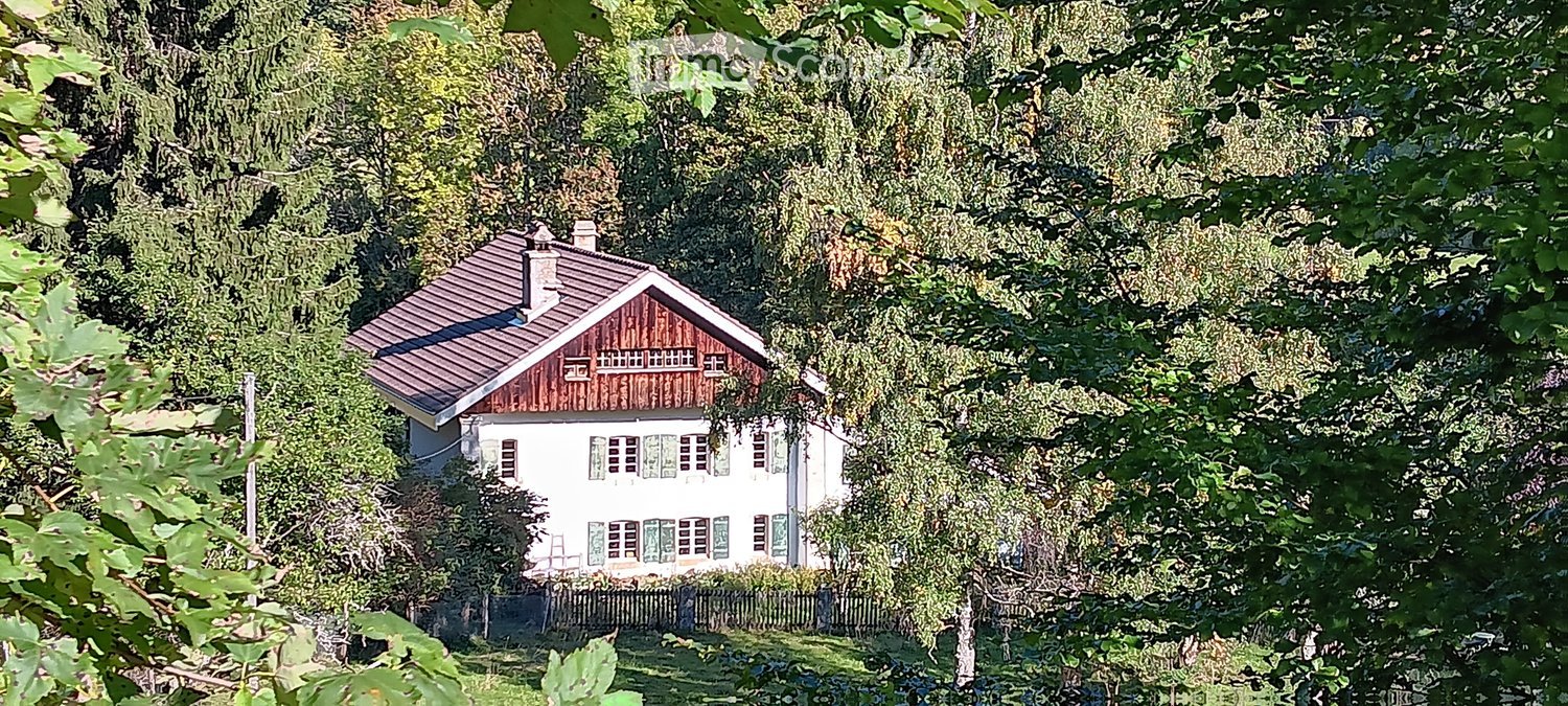 A two-story house with a red wooden roof and white walls, surrounded by lush green trees and foliage.