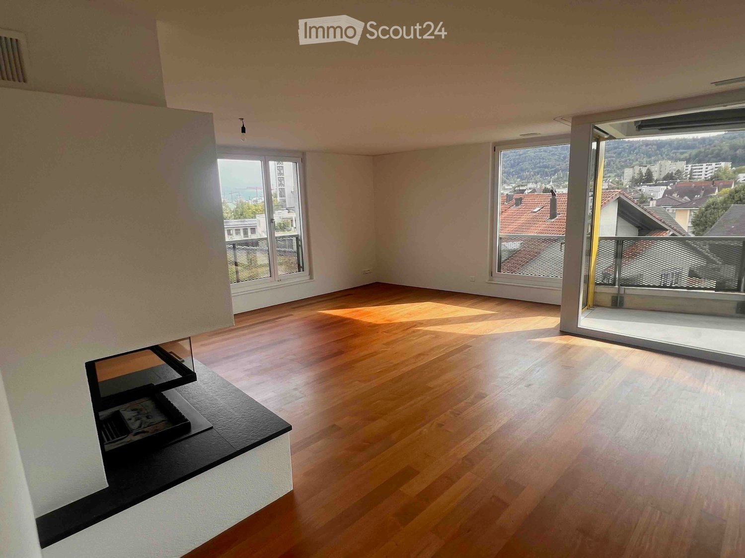 Empty room with wooden flooring, large windows, and a fireplace area. No furniture or equipment present.