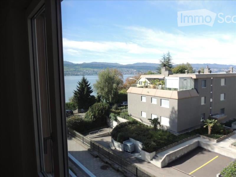 View from a window showing a modern multi-story building with a balcony, surrounded by greenery and overlooking a lake and distant mountains.