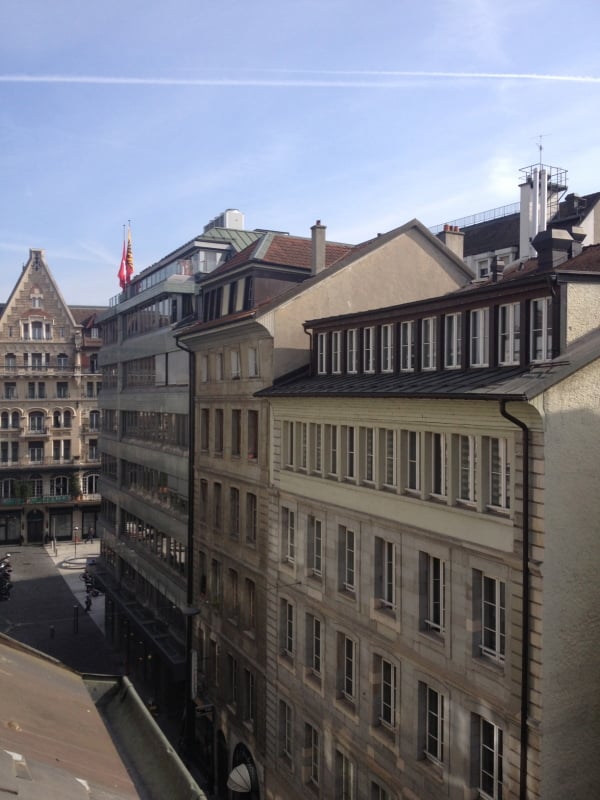 A view of a cityscape with multiple multi-story buildings, featuring classic architectural styles, chimneys, and a mix of flat and pitched roofs.