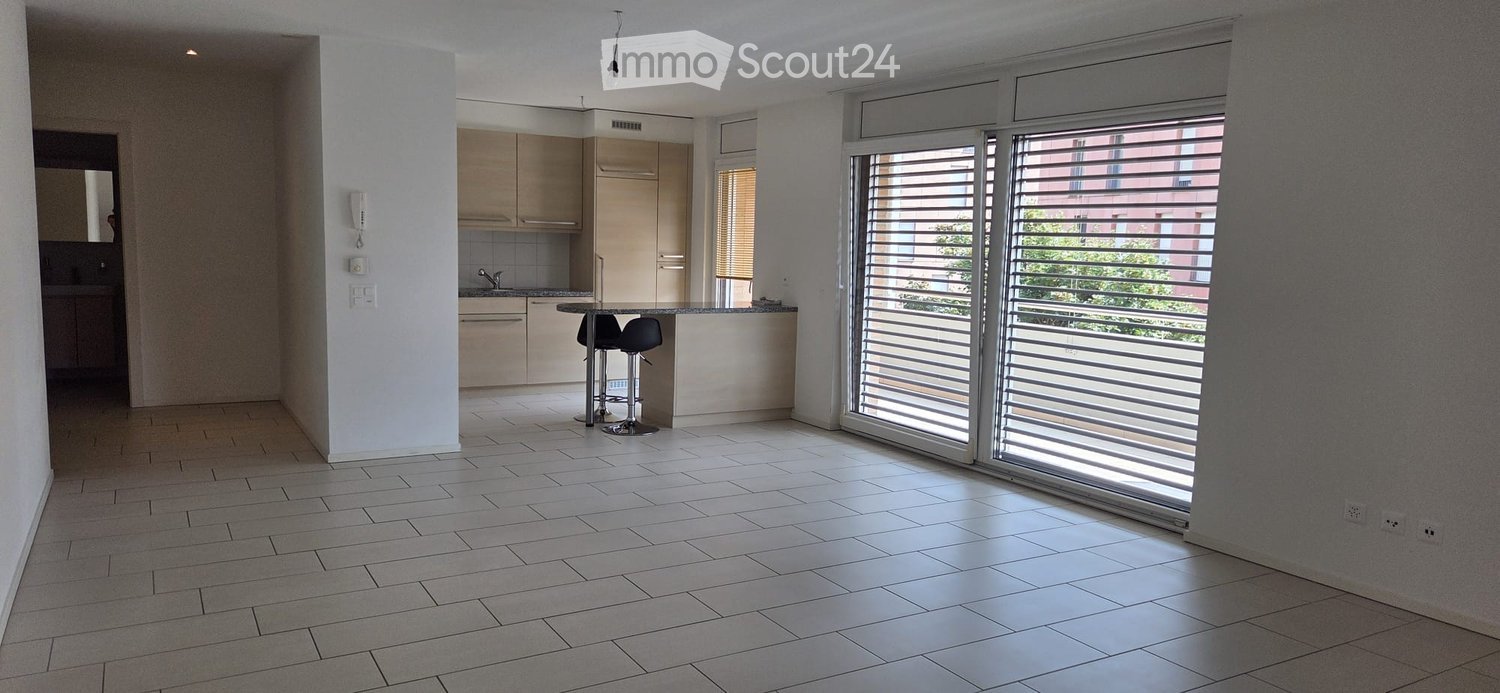 Bright, open-plan living and dining area with tiled flooring, large sliding glass doors leading to a balcony, and a small kitchen area with cabinets and a sink.