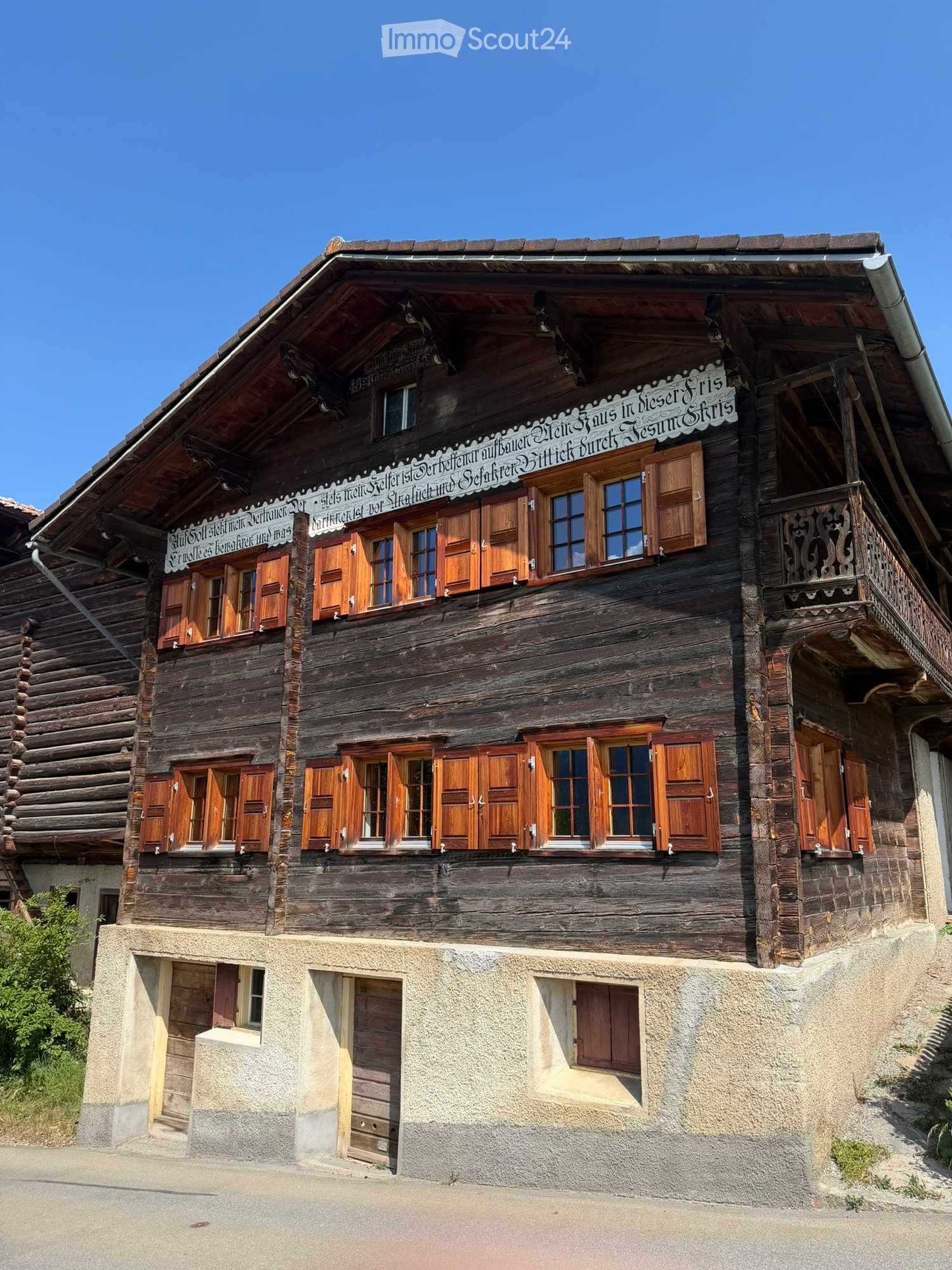 Traditional wooden house with multiple stories, wooden shutters, and a decorative balcony. The building has a steep, sloped roof and is situated on a raised ground floor.