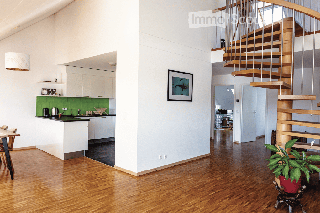 Open-plan living and kitchen area with light-colored cabinets, green backsplash, wooden flooring, a staircase with wooden steps and metal railings, and a dining table with chairs.