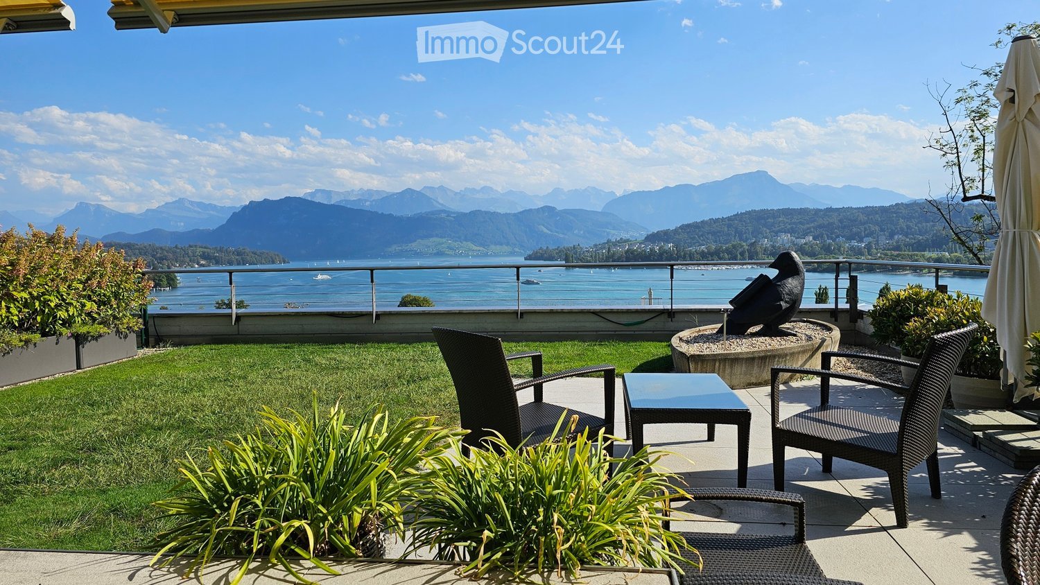 Balcony with a scenic view of a lake and mountains, featuring a small seating area with a table and chairs, surrounded by greenery and a decorative bird sculpture.