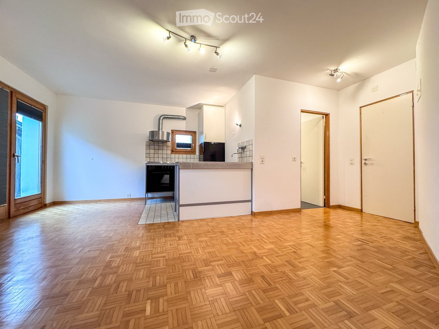 Empty kitchen with wooden herringbone flooring, white walls, and built-in appliances including a stove, oven, and microwave. The space features track lighting and a large window with blinds.