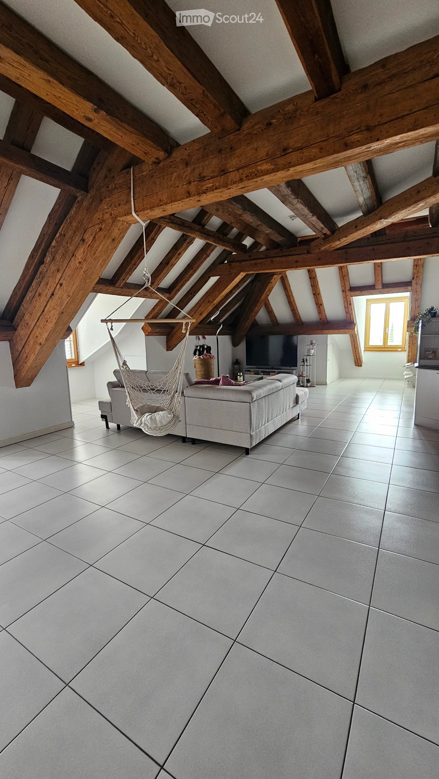 Loft-style living room with exposed wooden beams, tiled floor, grey sofa, hammock chair, television, small table, and shelves.