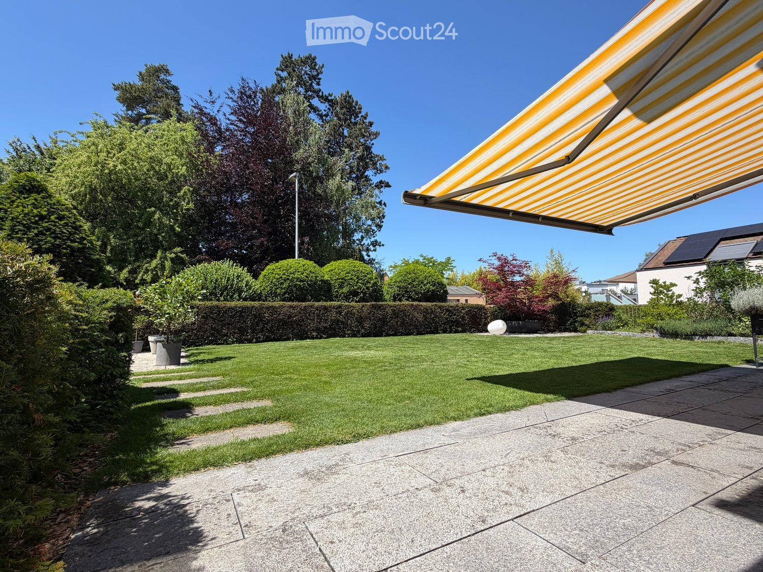 Well-maintained garden with a neatly trimmed lawn, a hedge border, potted plants, and a covered patio area with a striped awning.