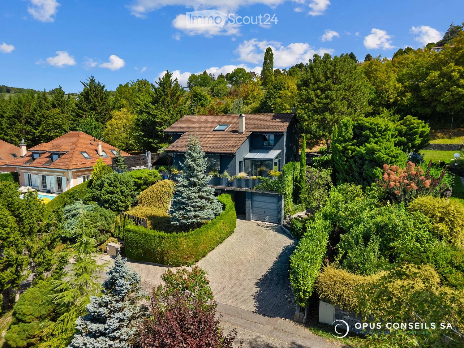 Aerial view of a two-story house with a red-tiled roof, surrounded by lush greenery, a driveway, a garage, and a swimming pool.