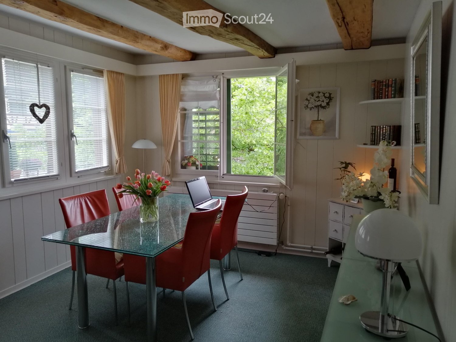 Dining table with 4 red chairs, glass table, flower vase, laptop, radiator, shelves, wooden beams on the ceiling