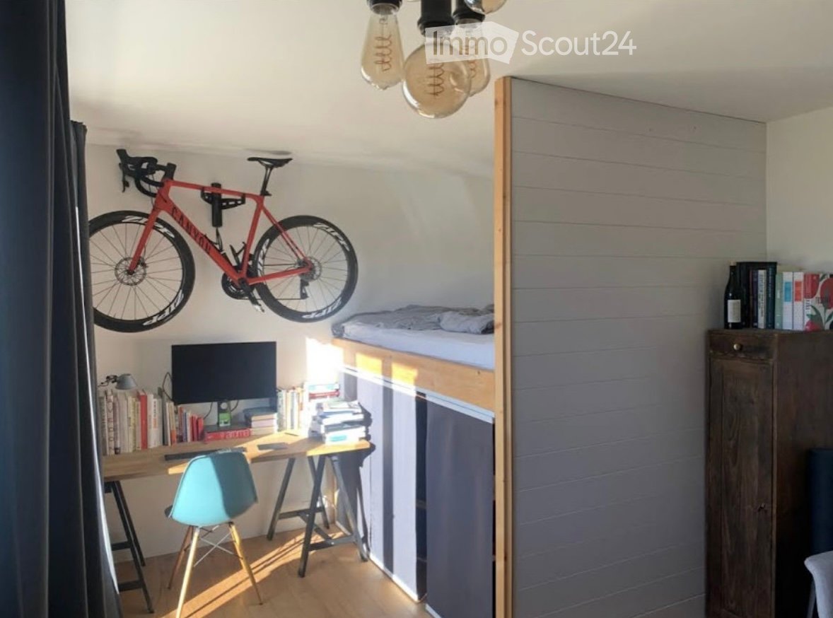 A compact, multi-functional room with a lofted bed, a desk with a computer screen, a bicycle mounted on the wall, and a small cabinet with books and a bottle.