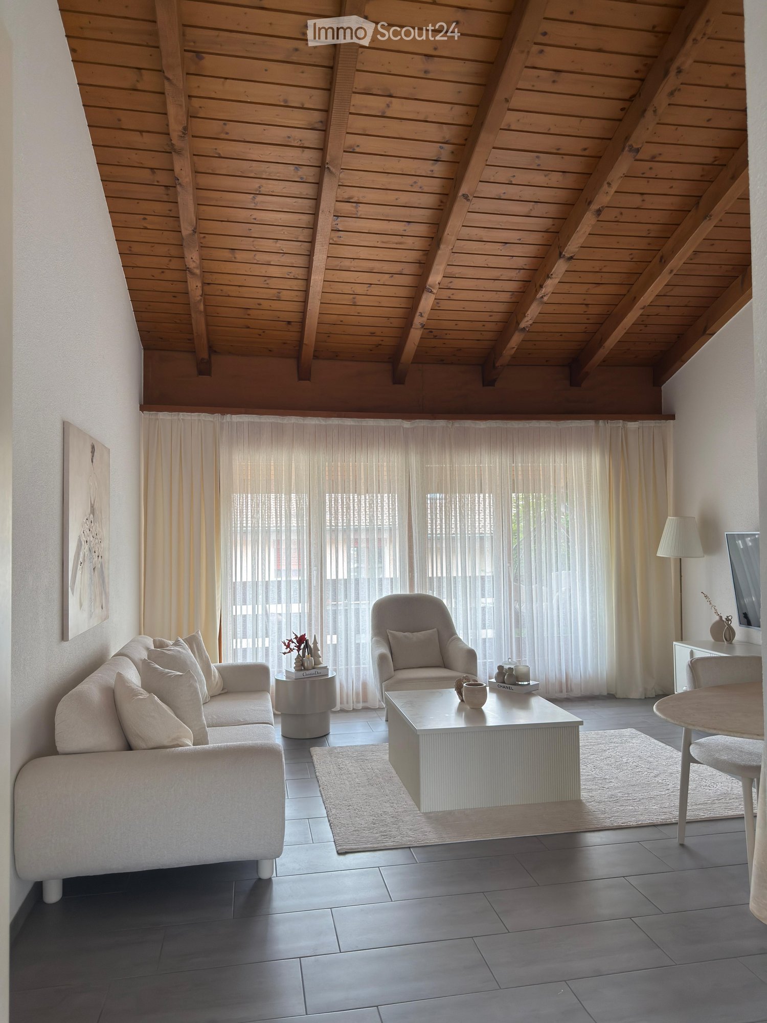 Modern living room with a grey sofa, armchair, coffee table, side table, and dining area with a table and chairs. The room features large windows with sheer curtains, a wooden slanted ceiling, and a neutral color palette.