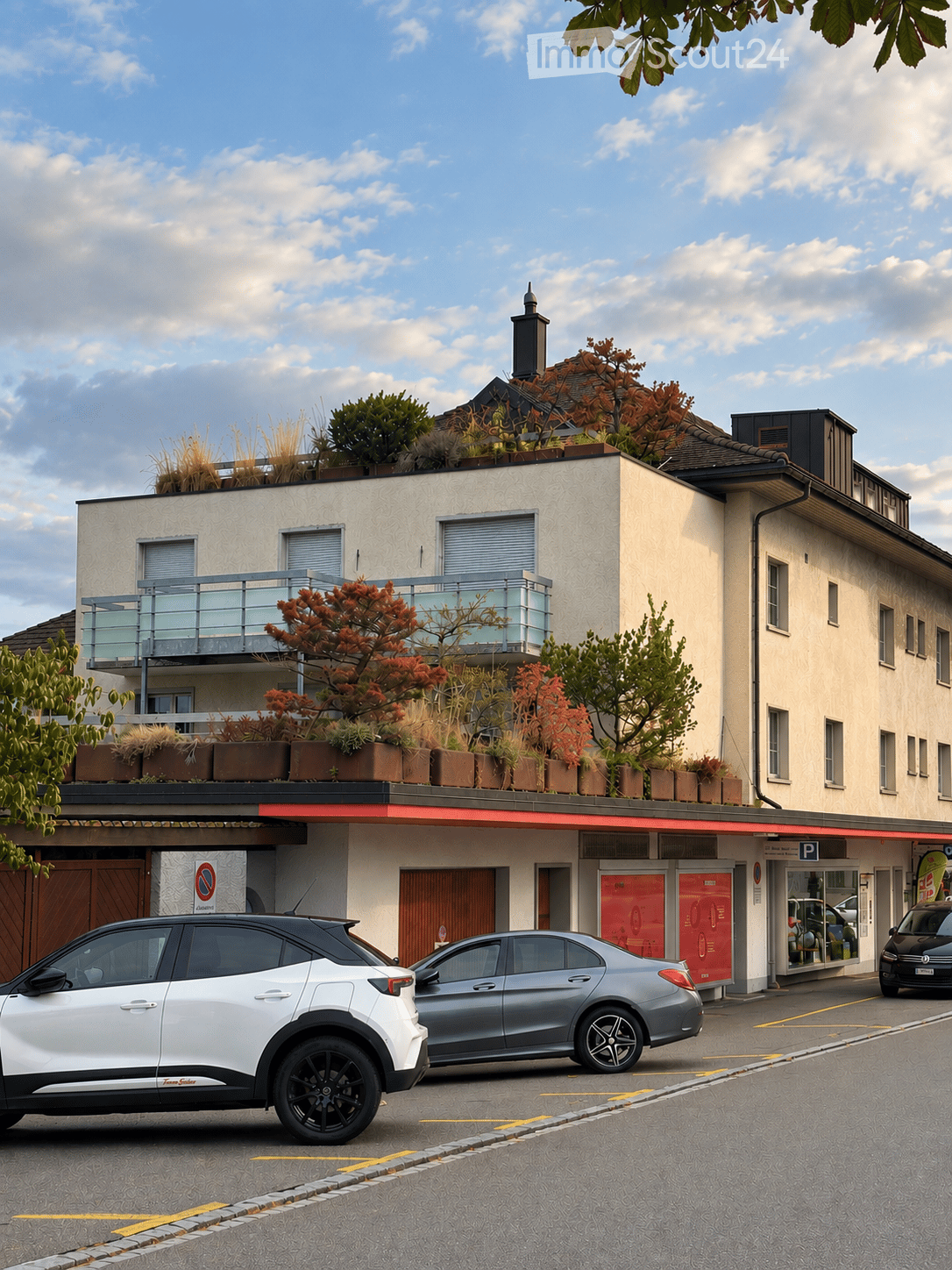 Modern multi-story building with a balcony, rooftop garden, and several parked cars in front.