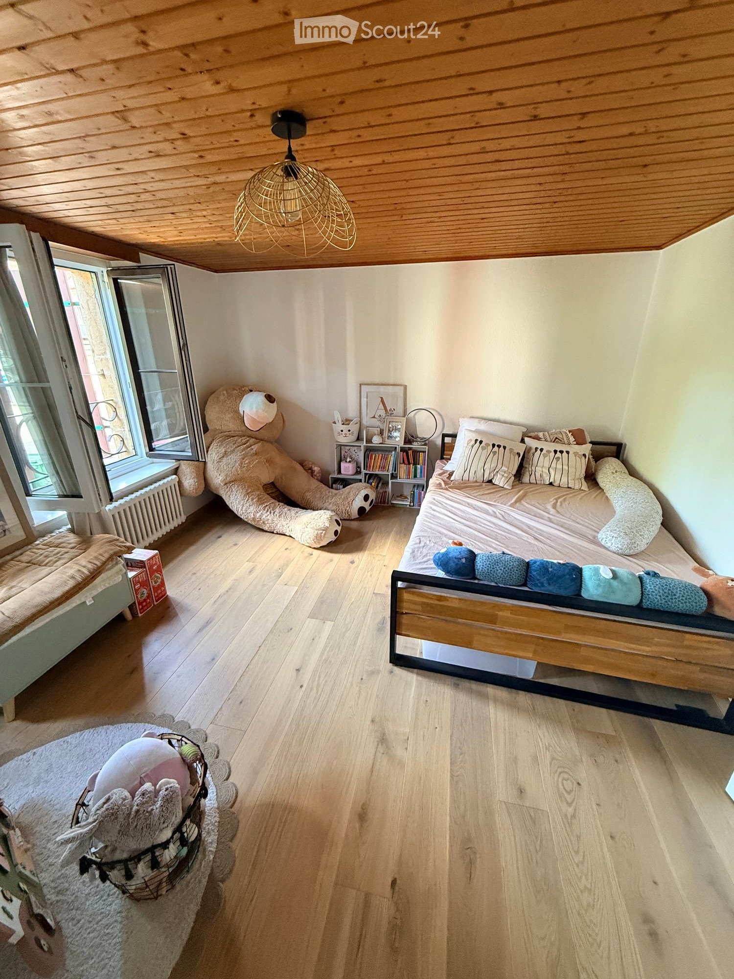 Cozy bedroom with wooden floors, slanted wooden ceiling, and large window. Features a bed with pillows, a large teddy bear, a bookshelf, and a basket with toys.