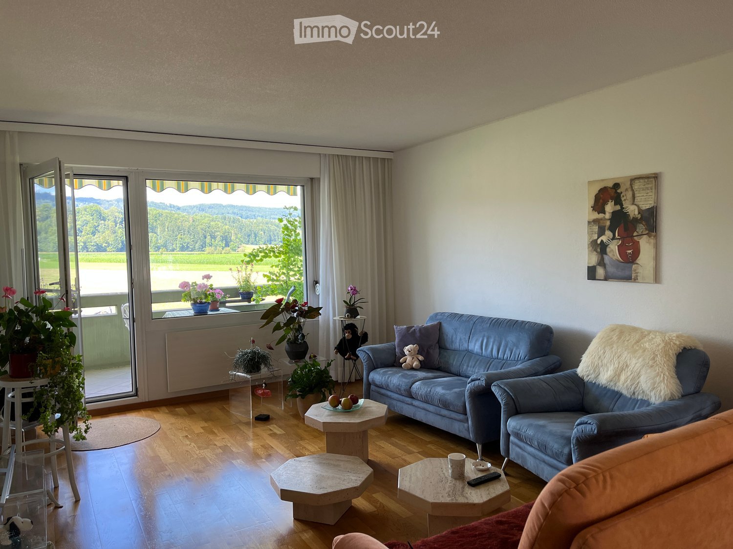 Bright living room with wooden flooring, large windows offering a scenic view, blue sofa, armchair, coffee tables, and decorative plants.