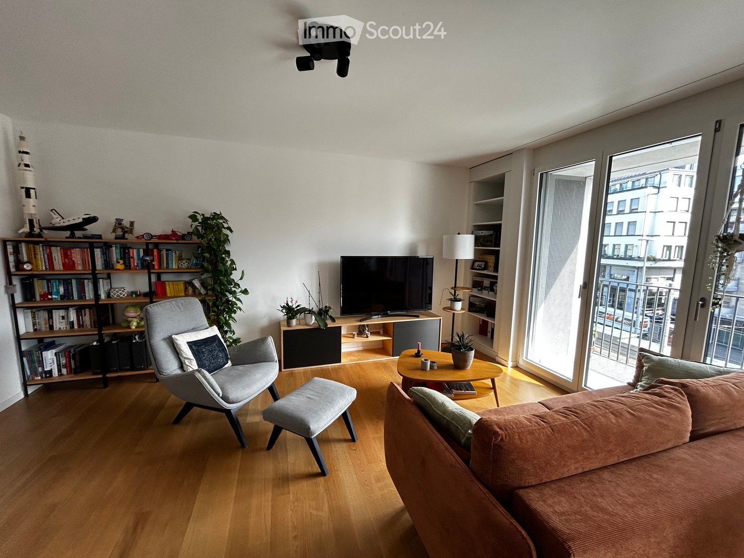 Modern living room with wooden flooring, large windows, a grey sofa, armchair, coffee table, bookshelf, and TV stand.