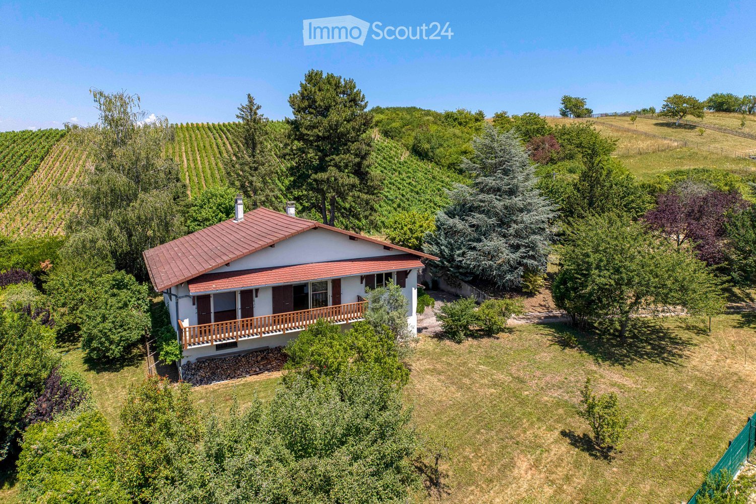 A two-story house with a red-tiled roof, white exterior, and a wooden balcony overlooking a vineyard and lush greenery.