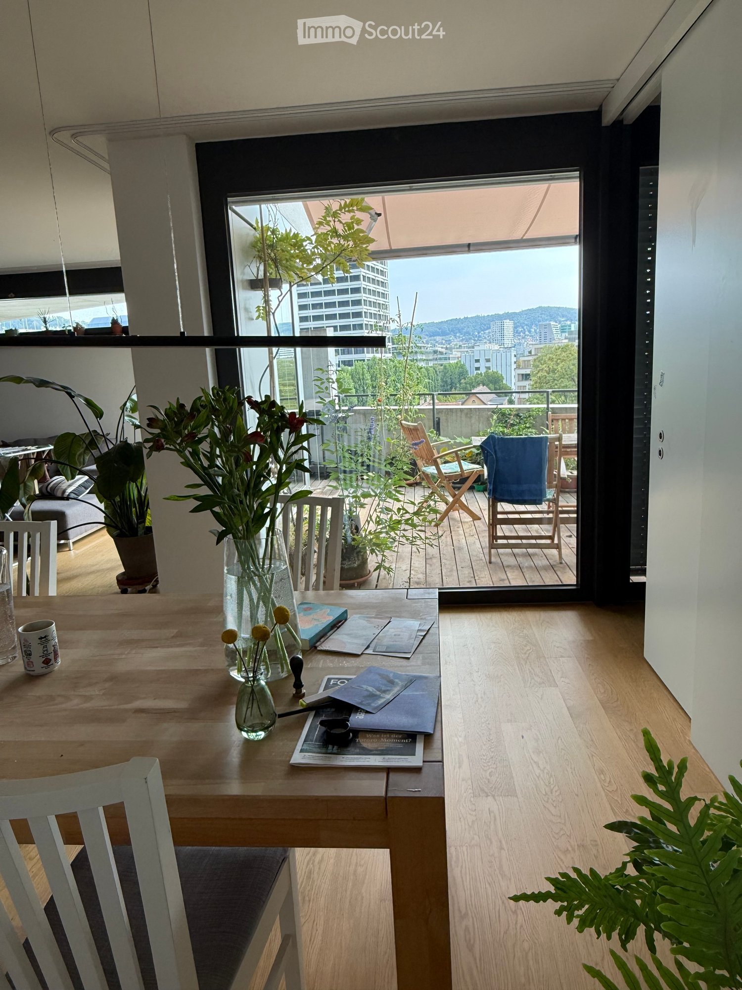 Bright room with wooden floor, large window offering city view, table with vase and cup, sofa, and outdoor seating area with chairs and plants.