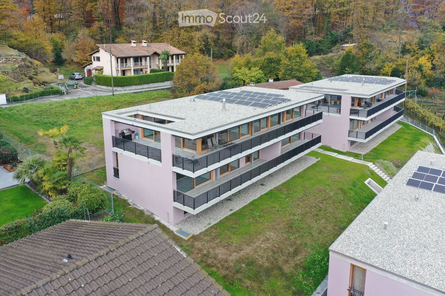 Aerial view of a modern, multi-story residential building with pink exterior, large windows, and multiple balconies. The roof features solar panels, and the surrounding area includes well-maintained lawns, trees, and neighboring houses.
