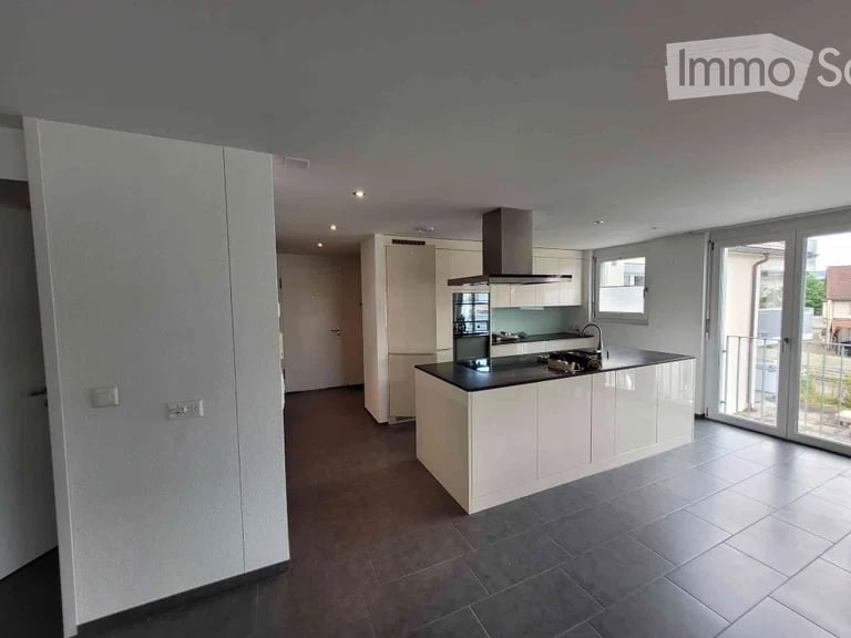 Modern, unfurnished kitchen with white cabinetry, a central island, and large windows leading to a balcony. The space features a sleek, minimalist design with a dark countertop and built-in appliances.