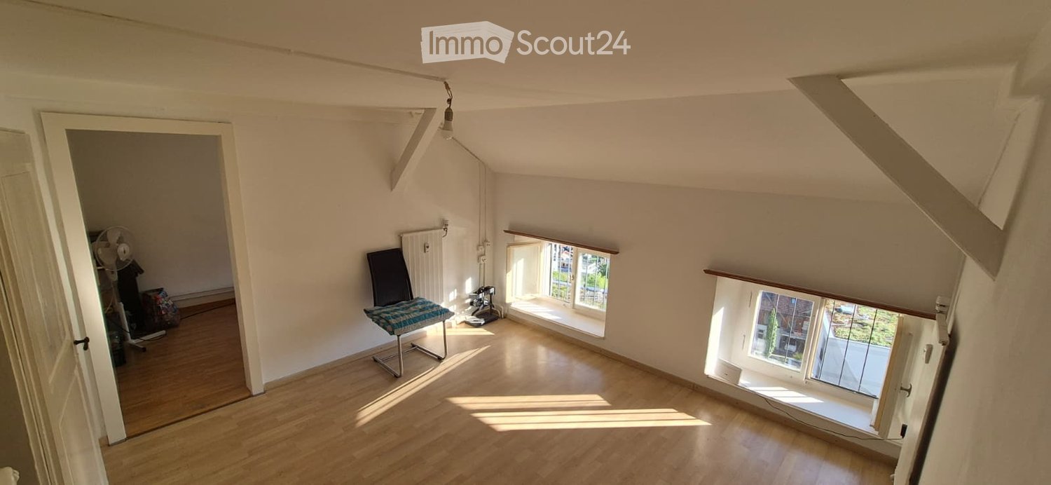 Bright attic room with wooden flooring, large windows, a single chair, a fan, and a radiator.