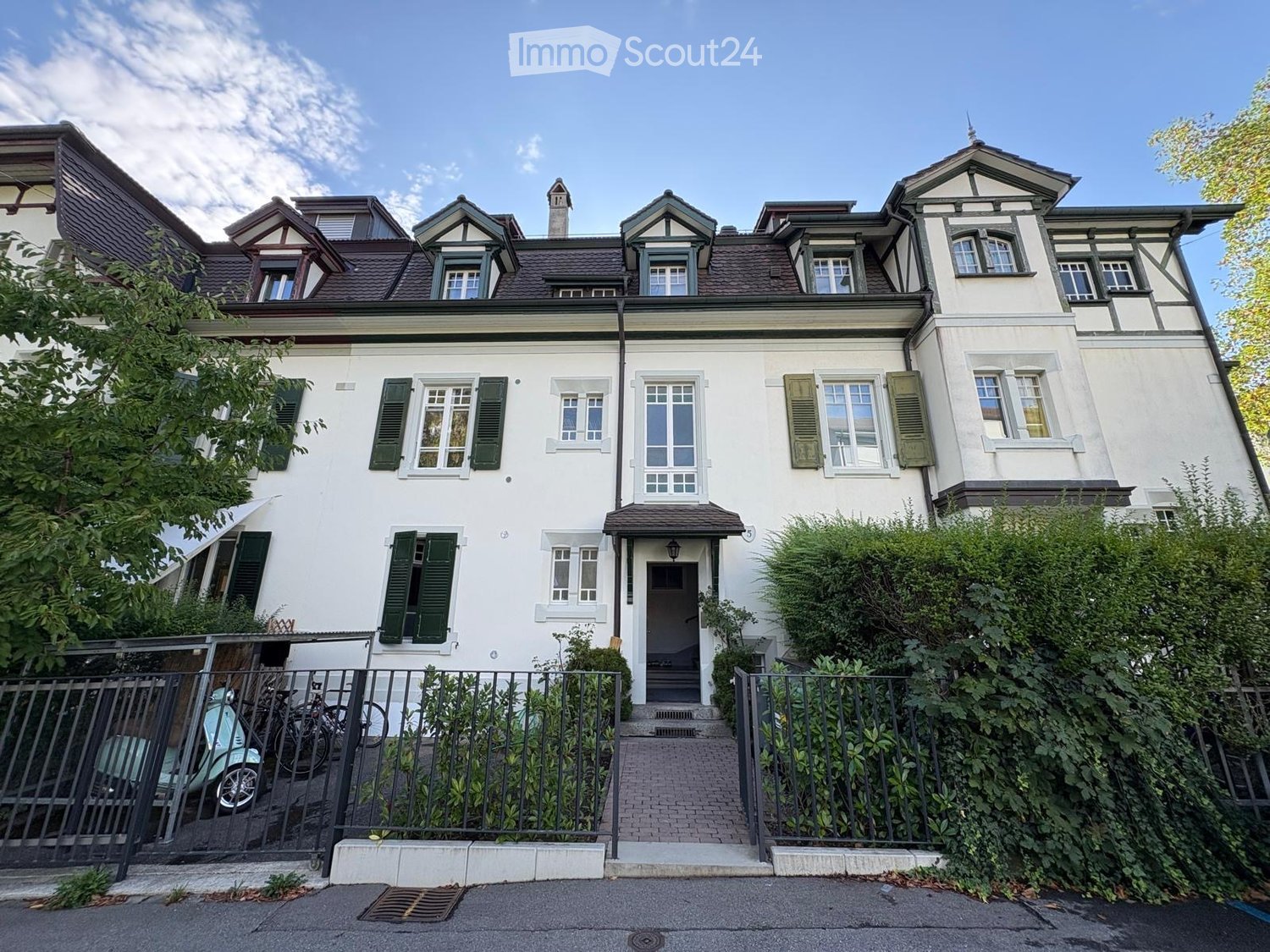 A two-story white house with green shutters, multiple windows, and a small entrance porch. The house features a pitched roof with dormer windows and is surrounded by a metal fence and greenery.