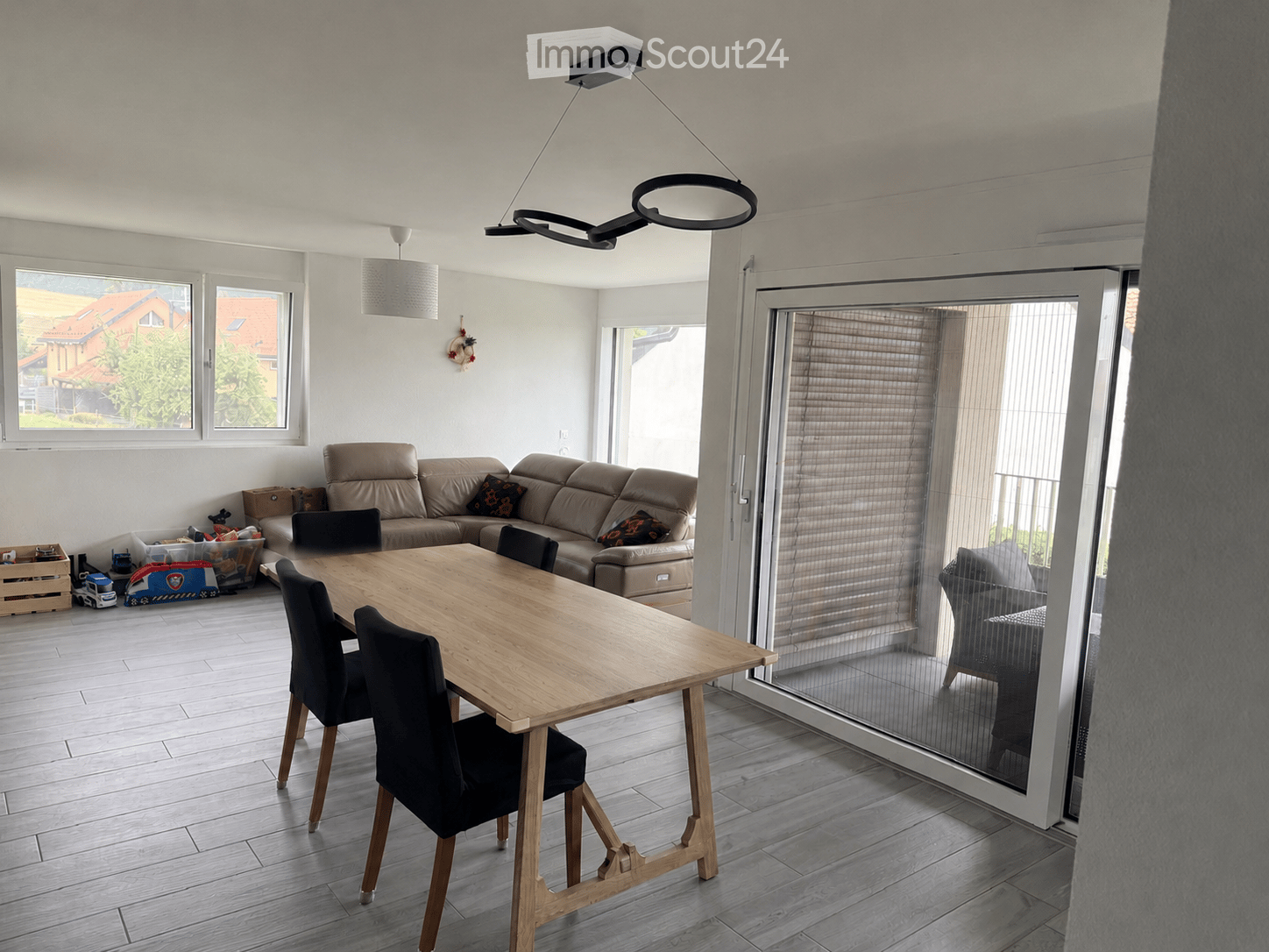 Bright living and dining area with a beige sofa, wooden dining table, black chairs, and a large window with blinds.