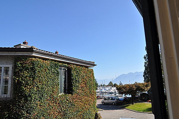 A white house with ivy-covered walls, overlooking a harbor with boats and distant mountains under a clear blue sky.