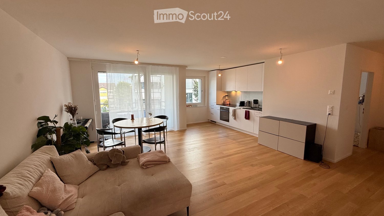 A modern, open-plan living and dining area with a sofa, dining table, chairs, and a fitted kitchen with white cabinets and appliances.