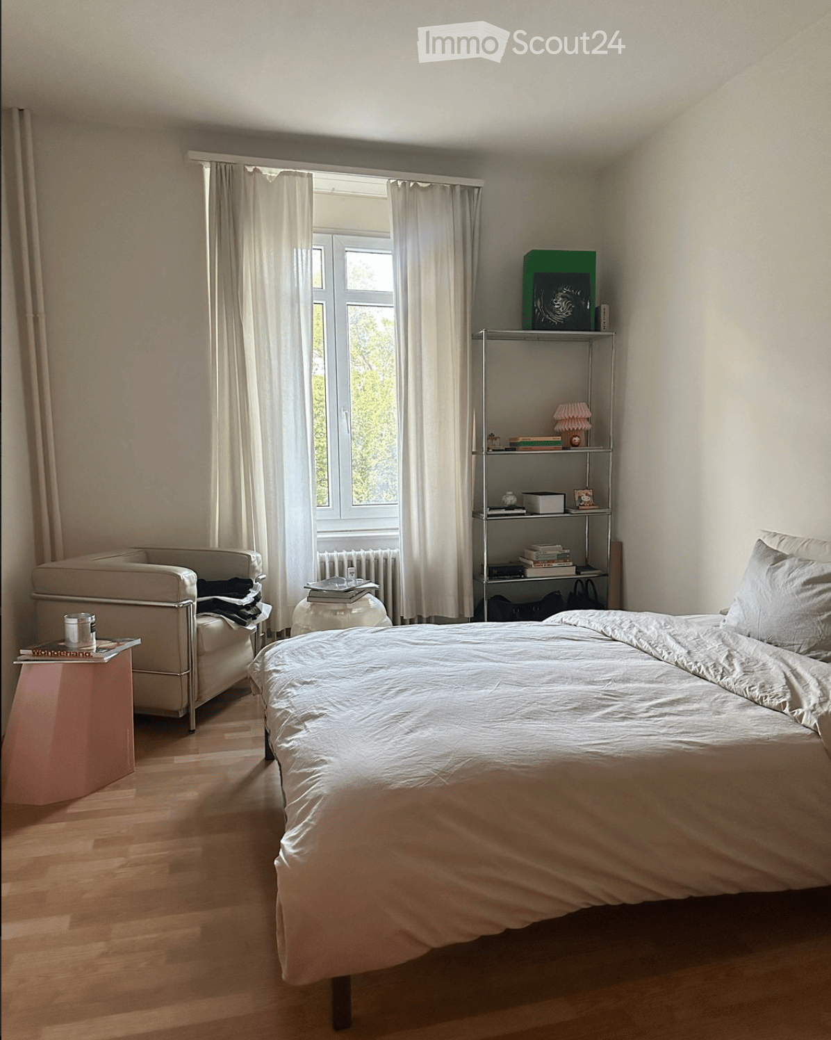 A well-lit bedroom with a large bed, a small sofa, a side table, and a metal shelf with books and decorative items. The room has wooden flooring and large windows with white curtains.