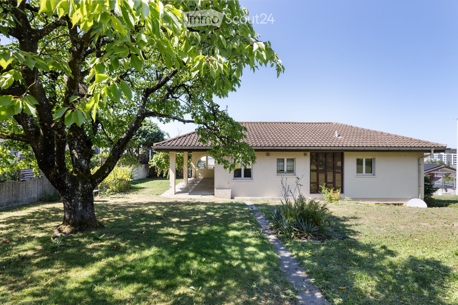 Single-story house with beige walls, tiled roof, and multiple windows. A covered entrance area is visible, surrounded by a well-maintained lawn and a large tree.