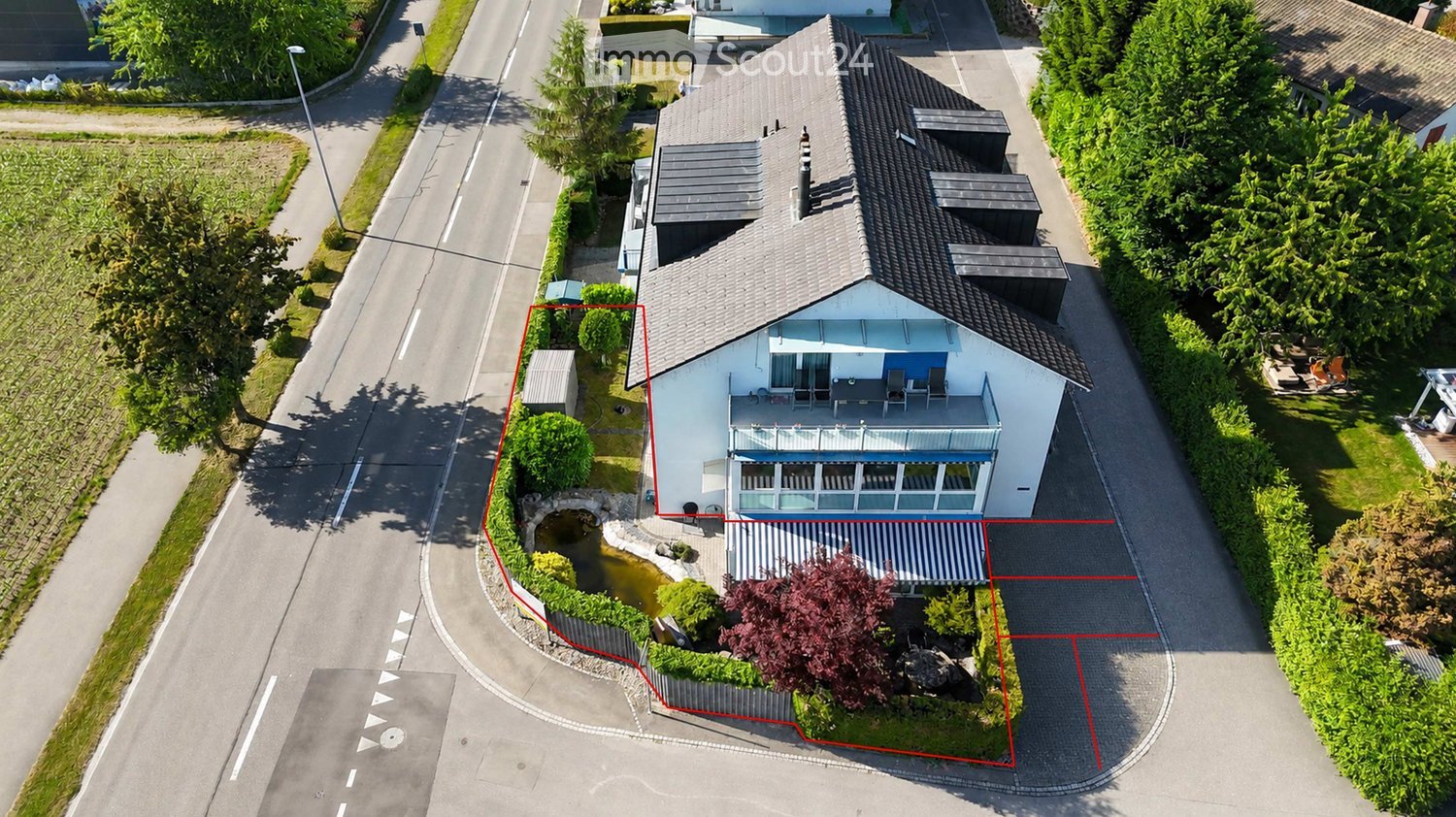 Aerial view of a two-story house with a modern design, featuring a balcony, a small pond in the front yard, and surrounding greenery.