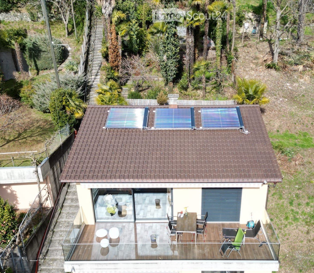 A two-story house with a tiled roof, solar panels, and a spacious balcony with outdoor furniture including a table, chairs, and a lounge chair.