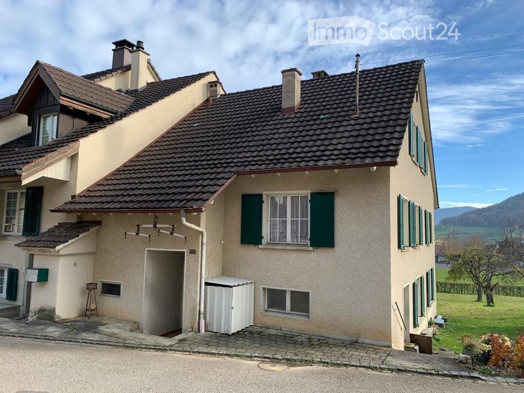Two-story house with beige walls, dark tiled roof, green shutters, and multiple chimneys. The house features a small garage and is situated in a rural area with a scenic view of the countryside.
