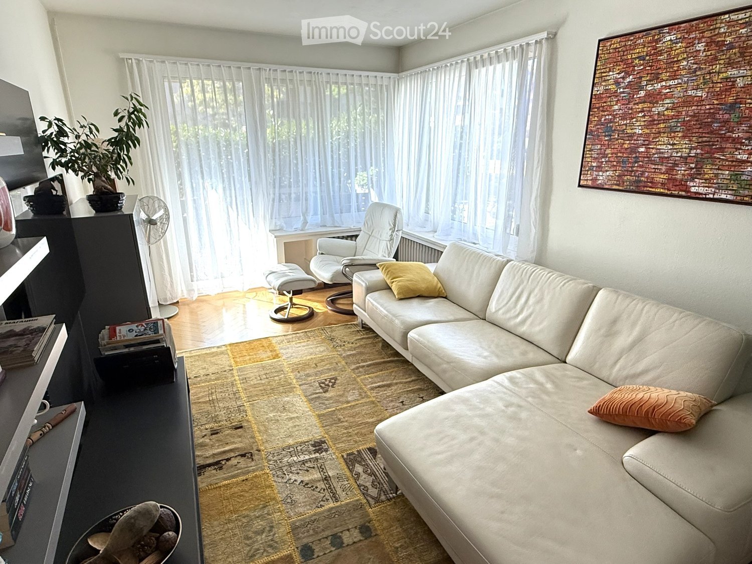 Modern living room with a white leather sofa, coffee table, armchair, fan, plant, and artwork on the wall.