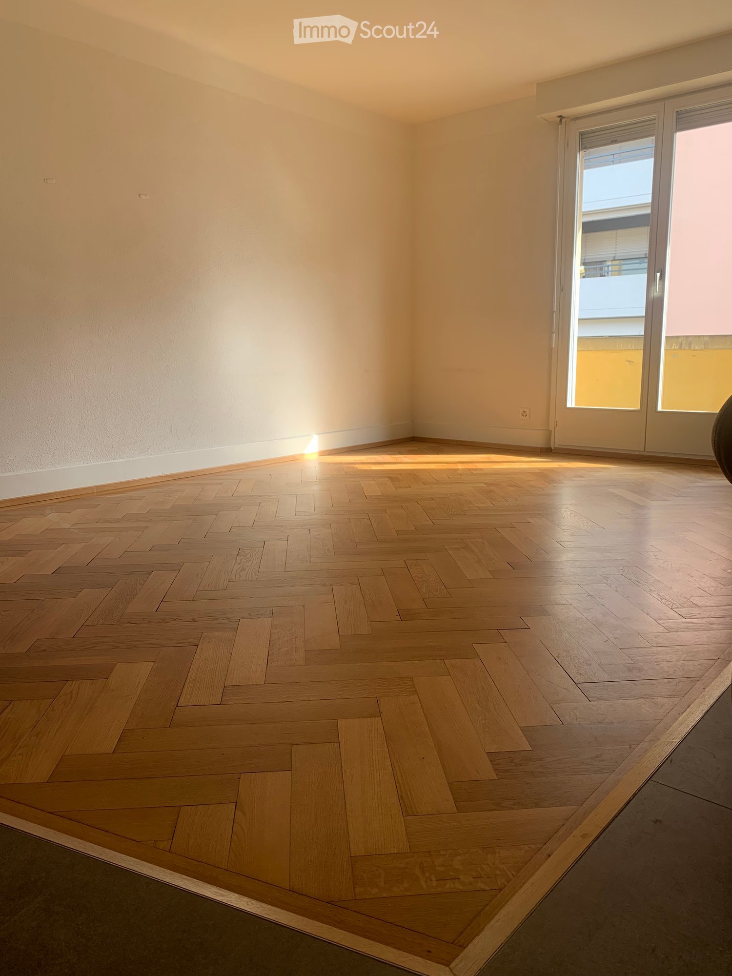 Empty room with light wooden herringbone flooring, white walls, and large glass doors leading to a balcony.