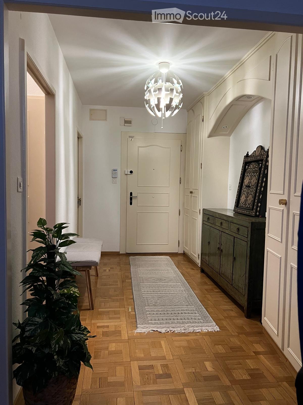 Hallway with wooden flooring, white walls, a modern chandelier, a small bench, a potted plant, a rug, and a cabinet with a framed artwork on top.