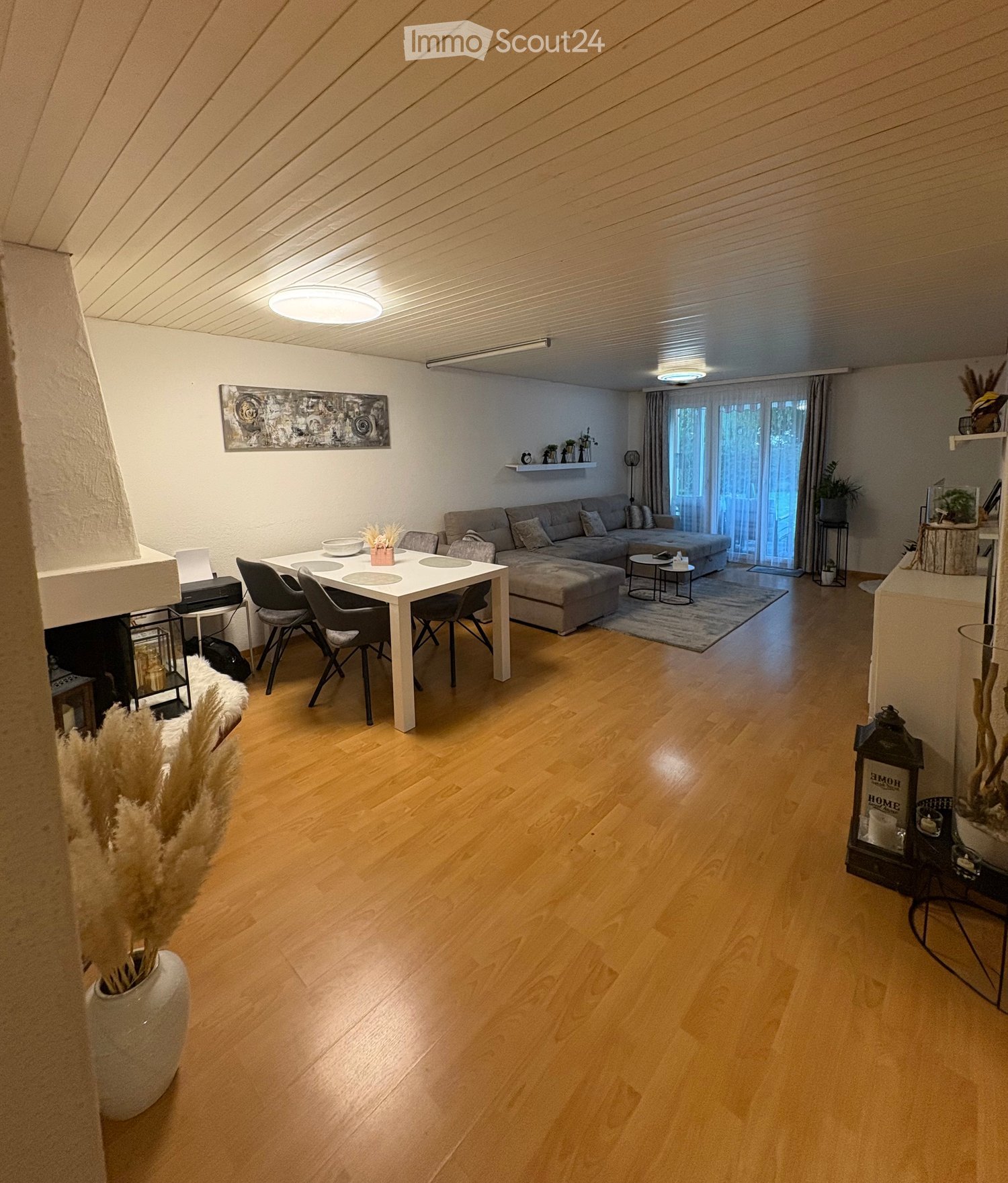 A modern living room with wooden flooring, a sectional sofa, a coffee table, a dining table with chairs, and a fireplace. The room features large windows with curtains and decorative elements like vases and artwork.