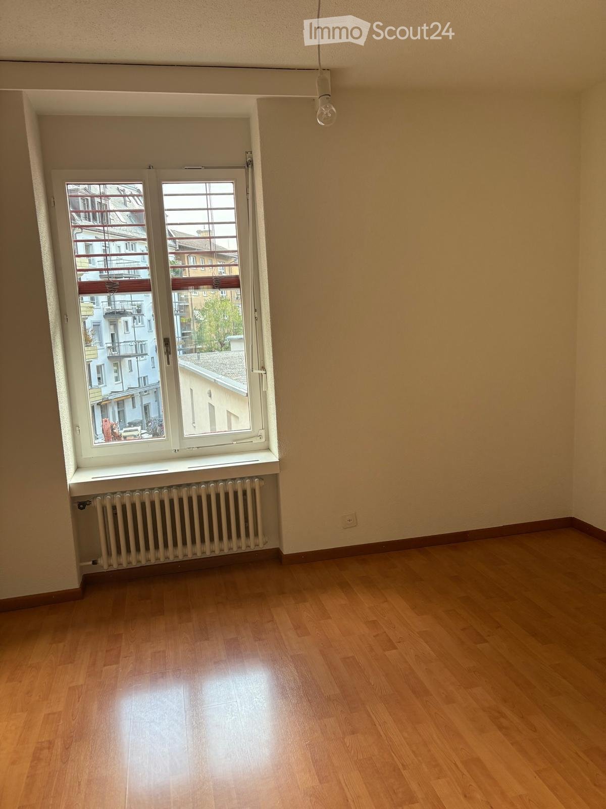 Empty room with wooden flooring, large window with radiator below, and visible cityscape outside.