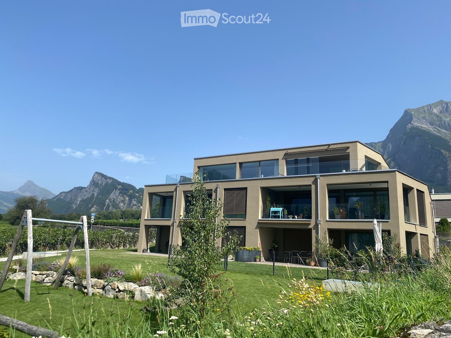 Modern two-story house with large windows and balconies, surrounded by a well-maintained lawn and garden, with mountains in the background.