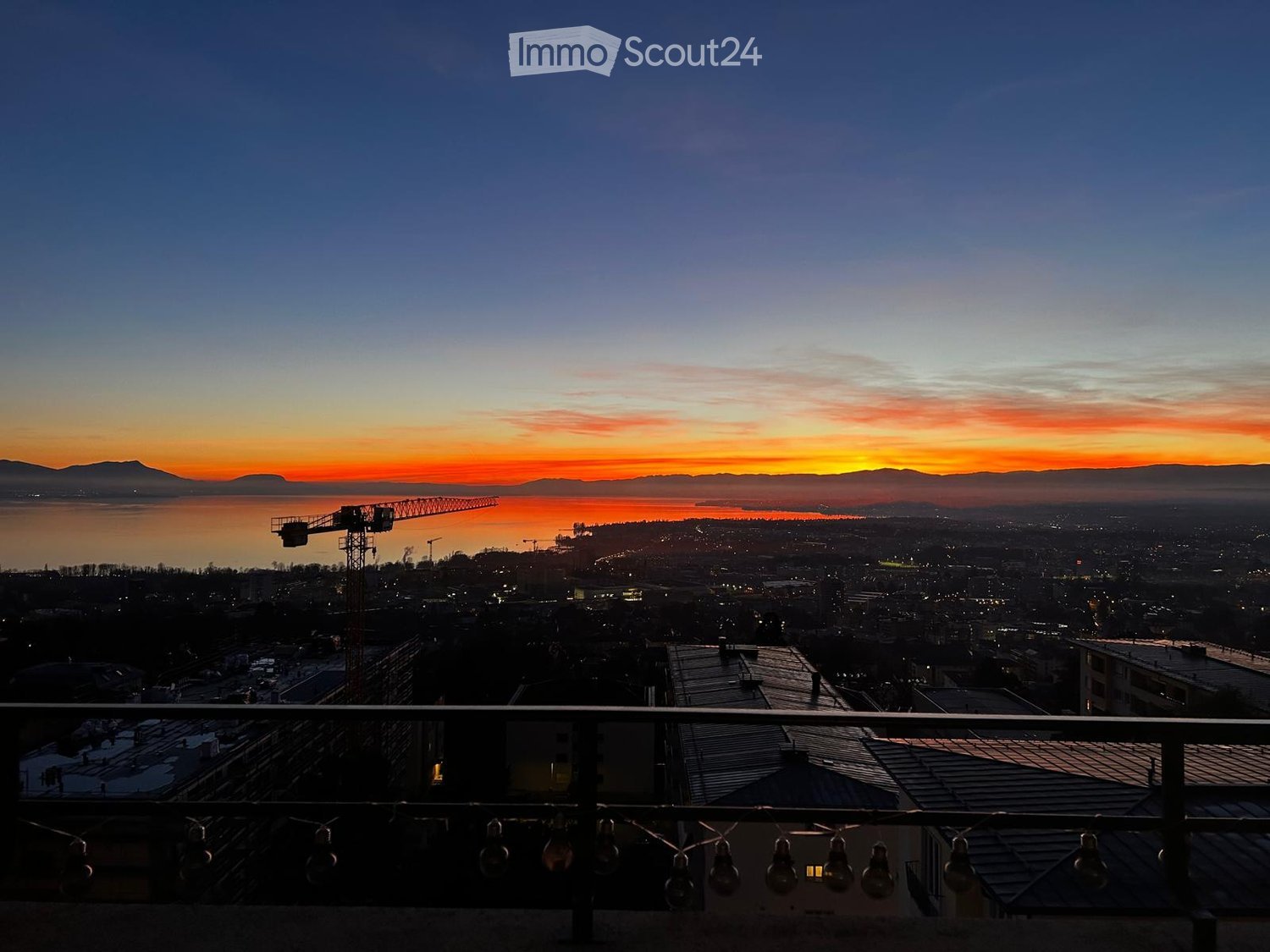 Balcony overlooking a cityscape at sunset, with a body of water and distant mountains visible.