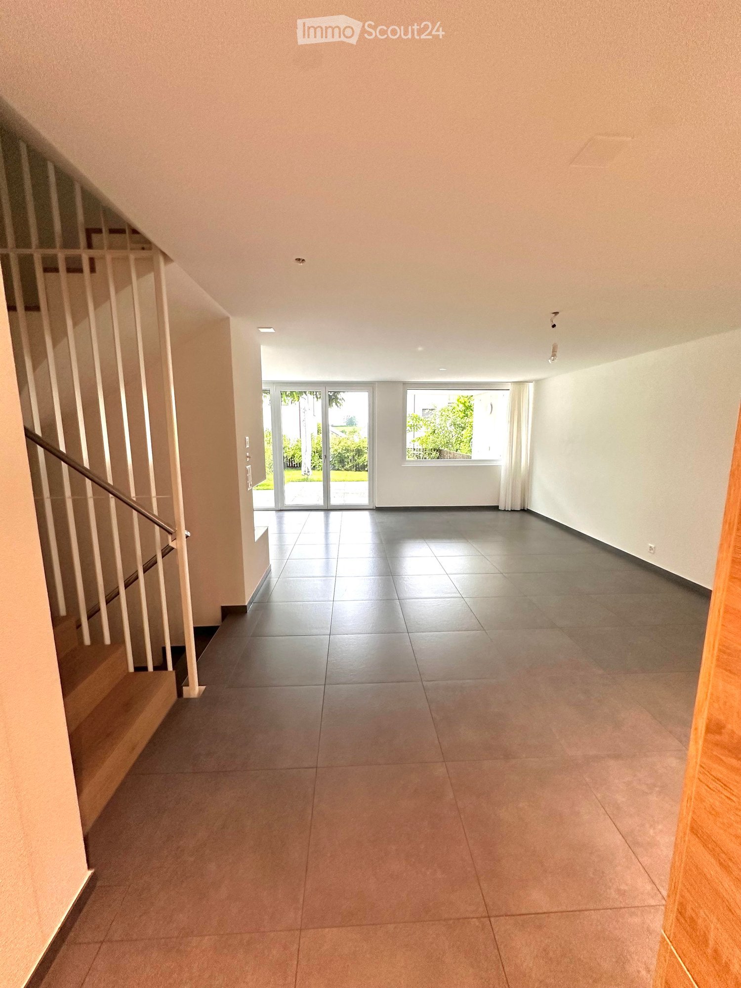 Empty hallway with tiled flooring, white walls, and a staircase leading upwards. Features include recessed ceiling lights, a window, and a sliding glass door leading outside.