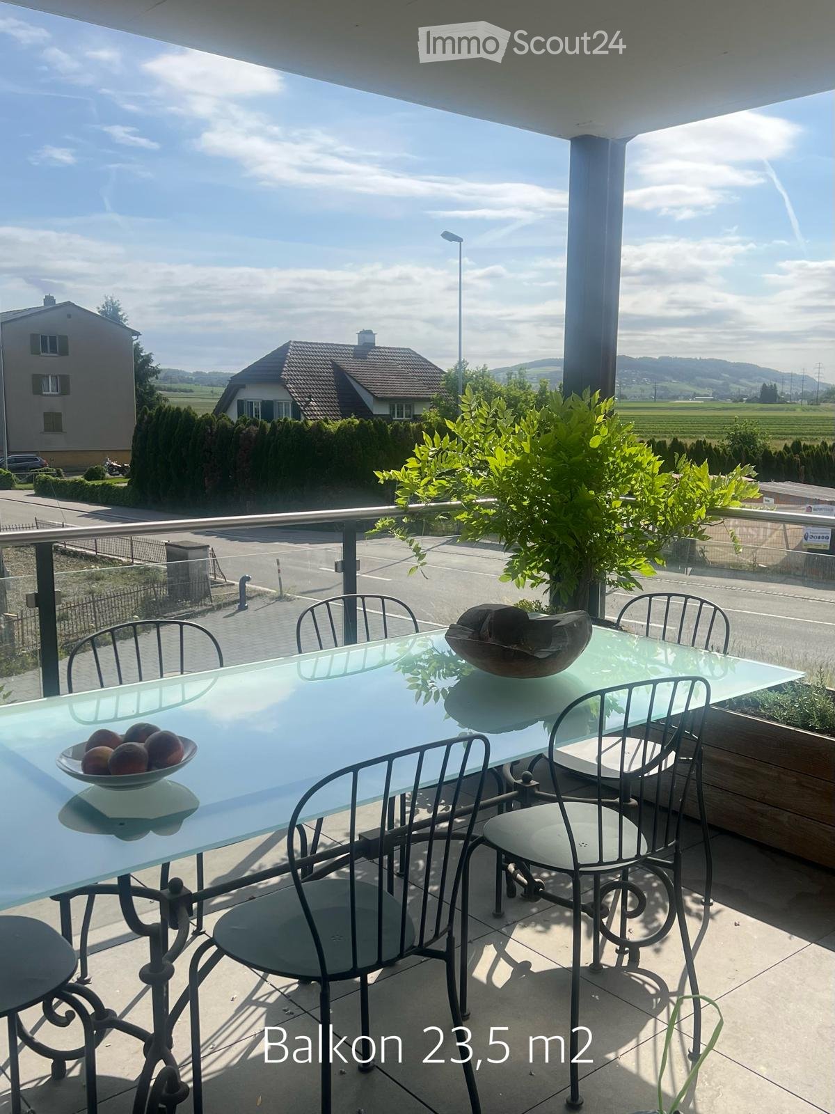 Balcony with a glass table, metal chairs, and a bowl of fruit, overlooking a scenic countryside view.