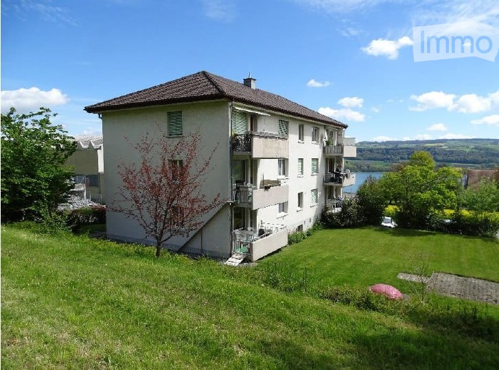 Multi-story residential building with balconies overlooking a scenic lake view, surrounded by green lawn and trees.