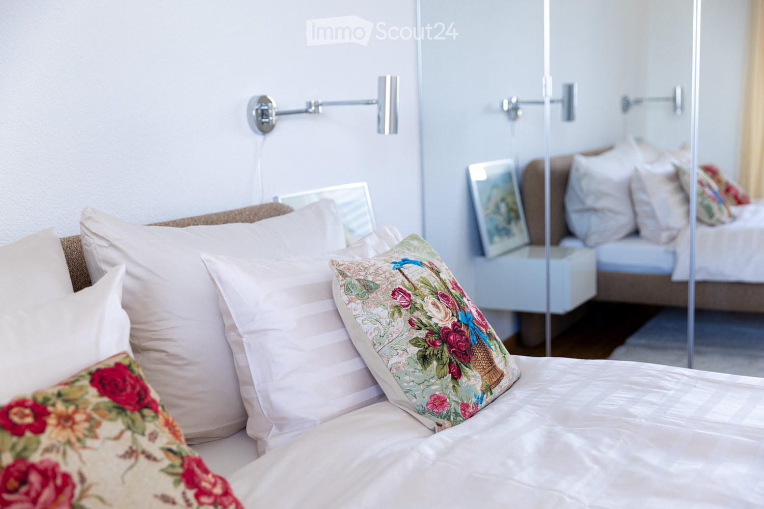 A well-lit bedroom with a neatly made bed, multiple pillows, a nightstand with a mirror, and a wall-mounted lamp.