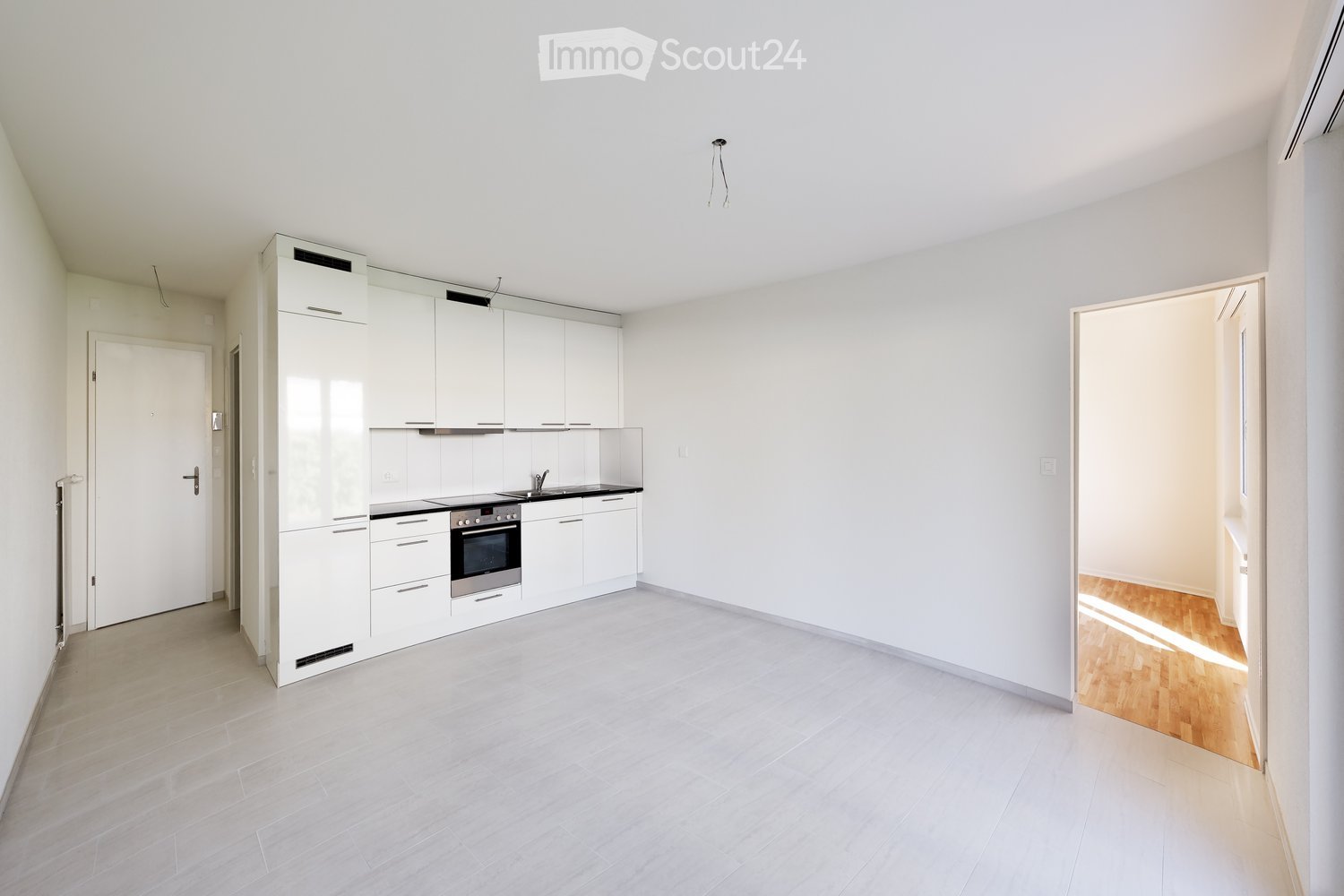 Modern, unfurnished kitchen with white cabinets, stainless steel appliances, and light grey tiles. The space is open and bright, with a doorway leading to another room with wooden flooring.