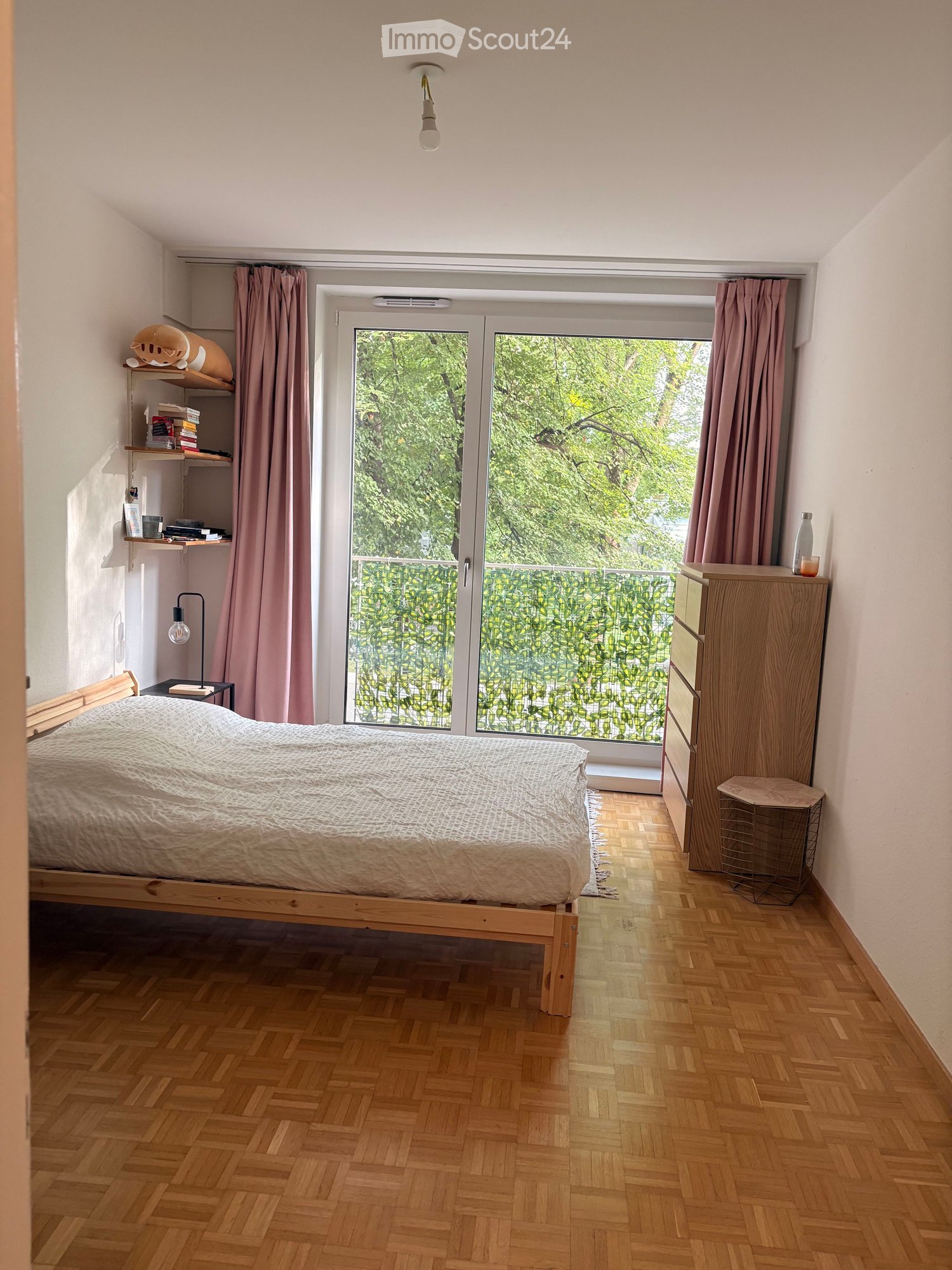 Cozy bedroom with wooden herringbone flooring, a single bed with a white duvet, a nightstand with a lamp, a shelf with books and decorative items, and a large window with pink curtains leading to a balcony.