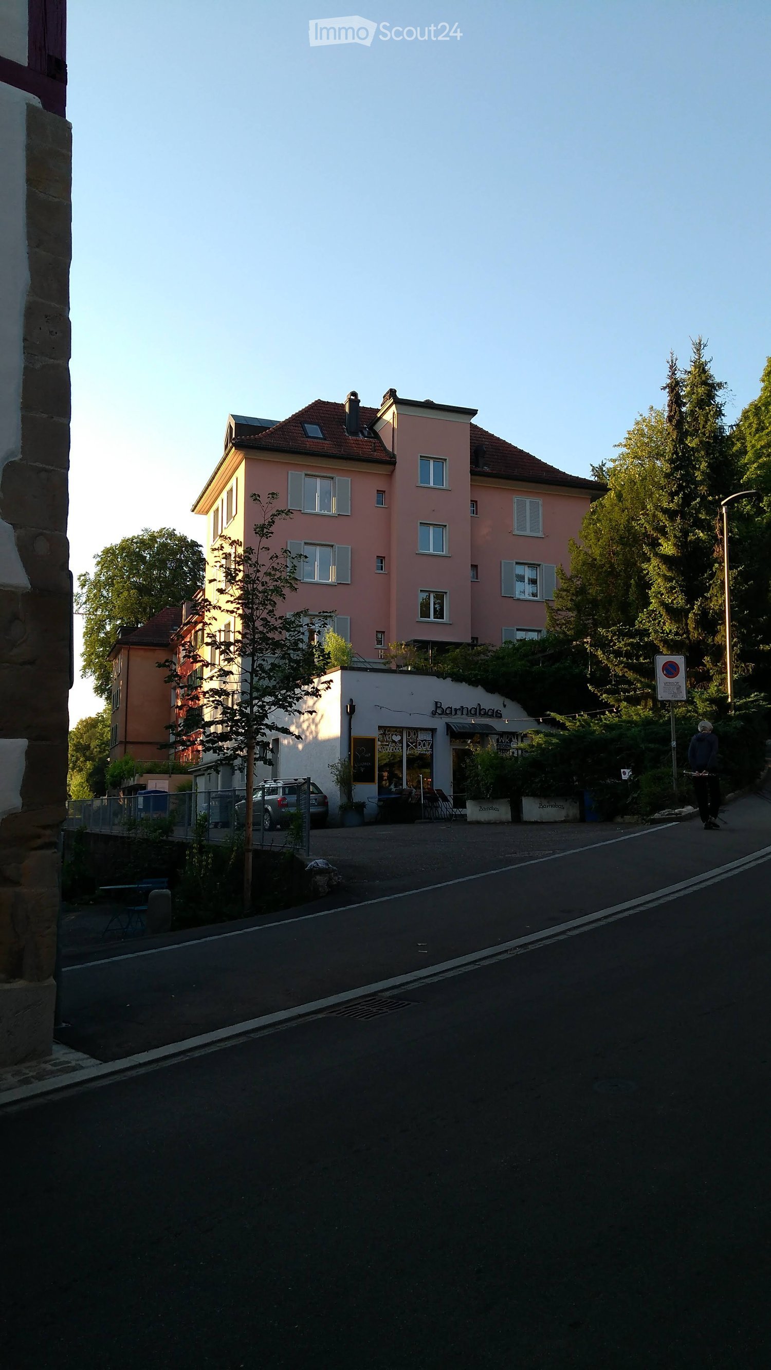 A multi-story residential building with pink facade and multiple windows, adjacent to a small shop with outdoor seating and a car parked nearby.