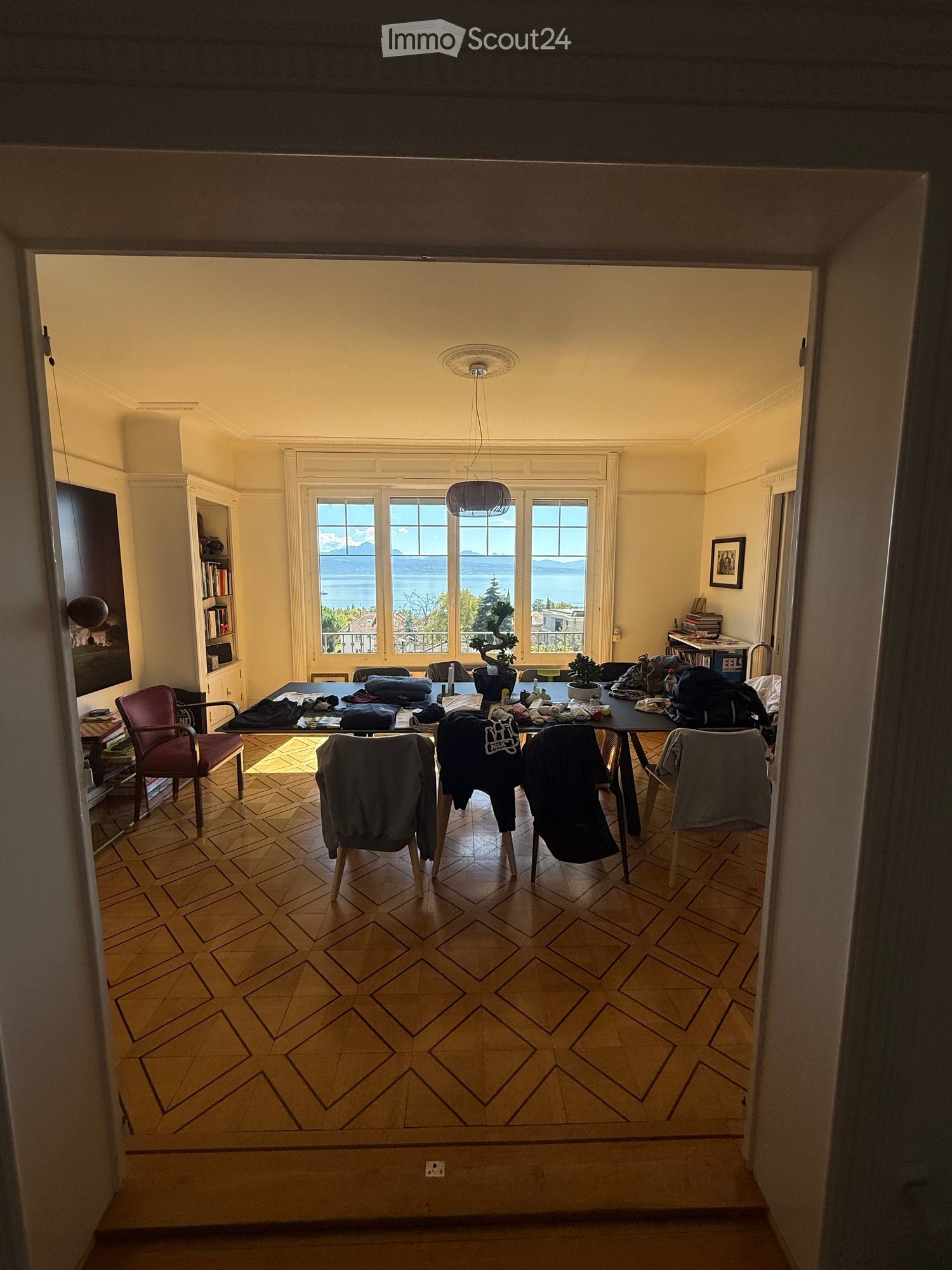 Bright room with wooden floor, large windows offering a scenic view, a table with chairs, potted plants, and a bookshelf.
